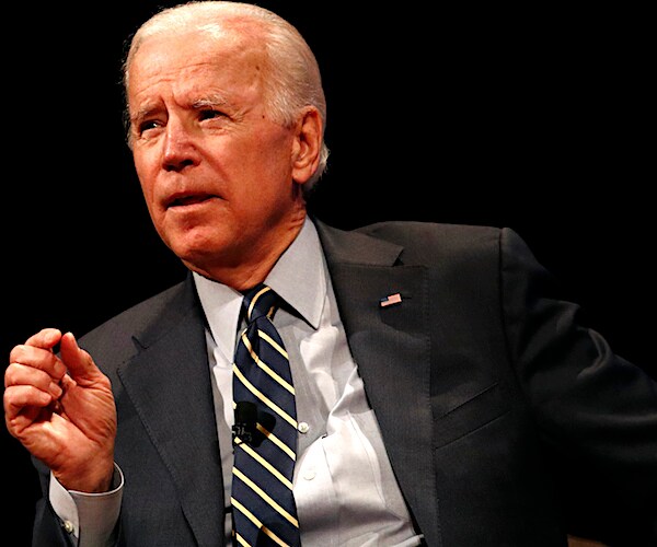 joe biden sits and gestures with his right hand while delivering his remarks on stage during a conference