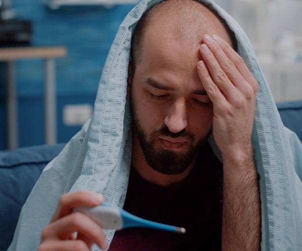 a man feeling sick with a blanket on his head, looking at thermometer