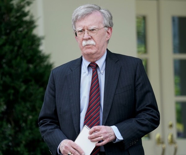 bolton in a dark gray suit and red and blue striped tie walking outside