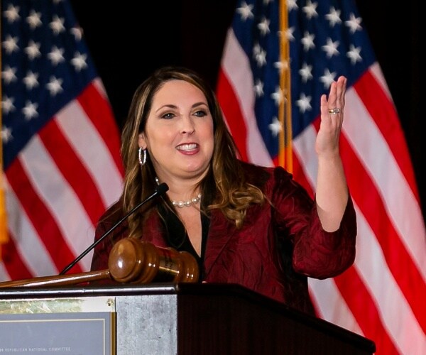 mcdaniel in a burgundy coat speaking at a podium in front of american flags