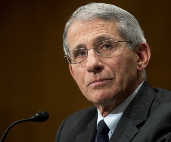 fauci in a dark gray suit and black tie sitting in front of a microphone