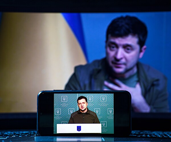 volodymyr zelenskyy speaks to his country on television as someone watches on a smart phone in front of the screen