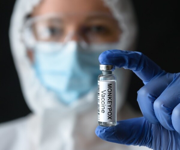 healthcare worker in protective gear holding a vial labeled 'monkeypox vaccine' up to camera