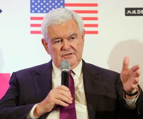 newt gingrich in a black suit and purple tie sitting down and speaking into a microphone