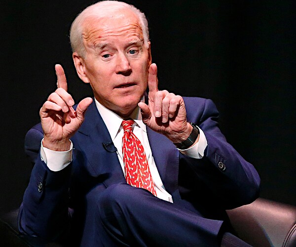 joe biden throws up both index fingers while seated and speaking on stage at a conference