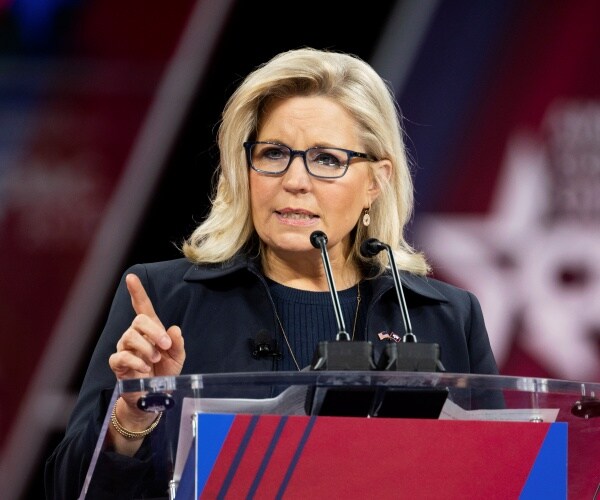 liz cheney in a dark blue top and black coat speaking at a podium at cpac