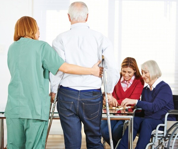 nurse in scrubs helps an old man in a shirt and jeans walk to a table with two women sitting down