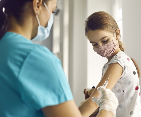 young girl holding teddy bear, wearing mask, getting a shot