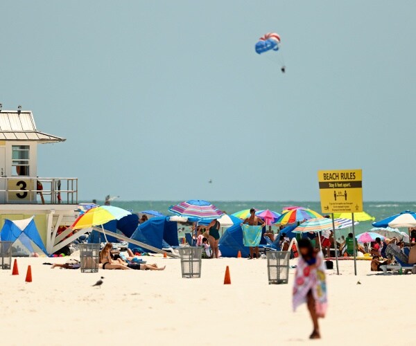 people sit under colorful umbrellas on the beach with a lifeguard watching and a sign telling people to social distance