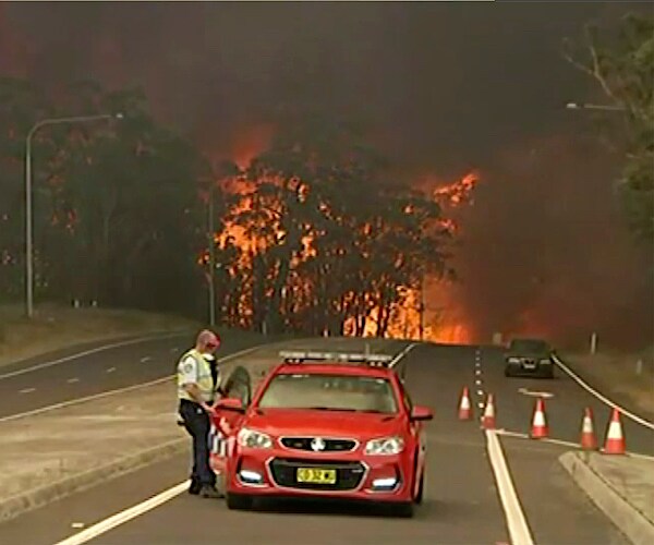 australian wildfires rage behind a police officer and a stopped car on a roadway