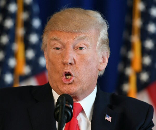 president donald trump is seen in a dark suit, red tie and white dress shirt with us flags in the background