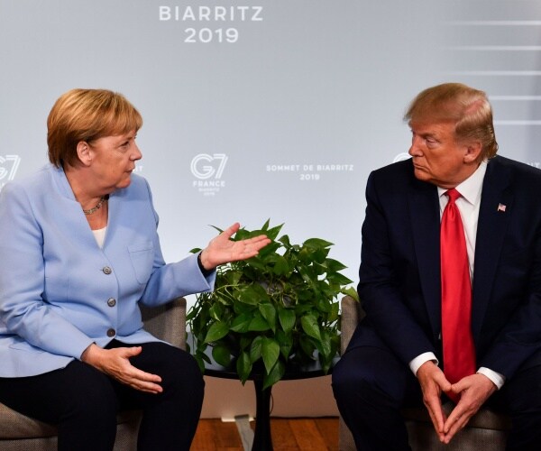 trump in a black suit and red tie sitting next to merkel in a light blue coat and black pants