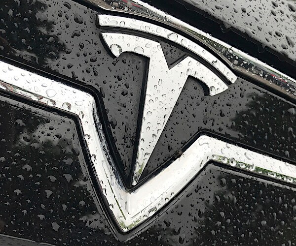 the telsa hood ornament with a silver tee on a black car with raindrops on it