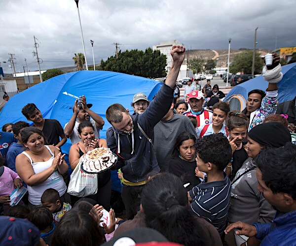 a group of caravan migrants stand around and a cheer each other's presence at the united states mexico border