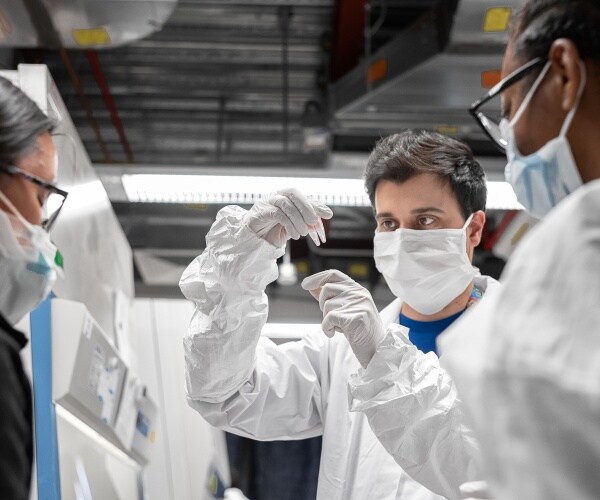 scientists in white protective suits and masks hold up small tubes with samples