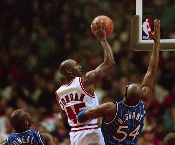 michael jordan in white jersey shooting a basketball into the hoop while horace grant in a blue jersey tries to block it