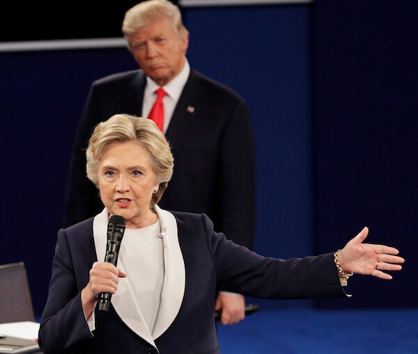 hillary clinton holds a mic in her right hand and extends her other one with donald trump lurking in the background