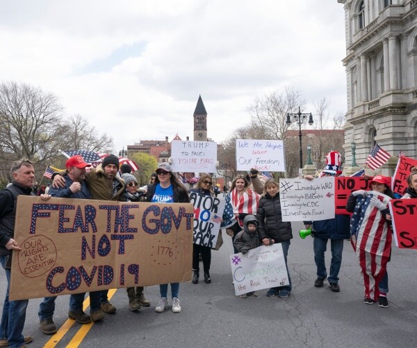 people hold up signs and american flags to protest lockdown with one saying "fear the gov't not covid19"