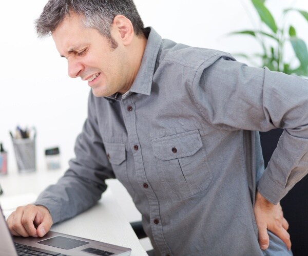 man at chair in front of desk with computer holding his lower back in pain