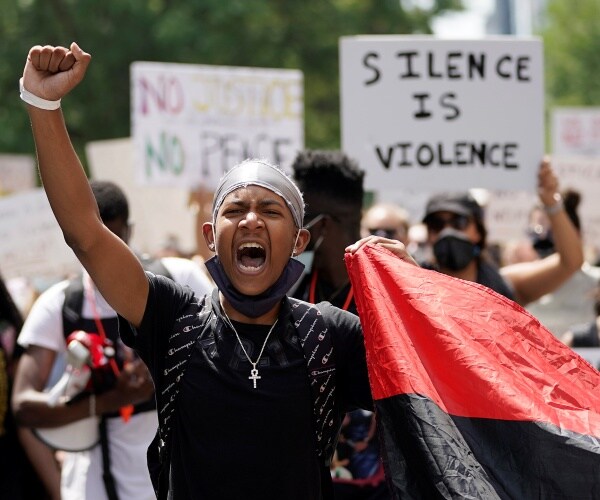 young man raises a fist with a crowd of protesters walking and holding signs behind him