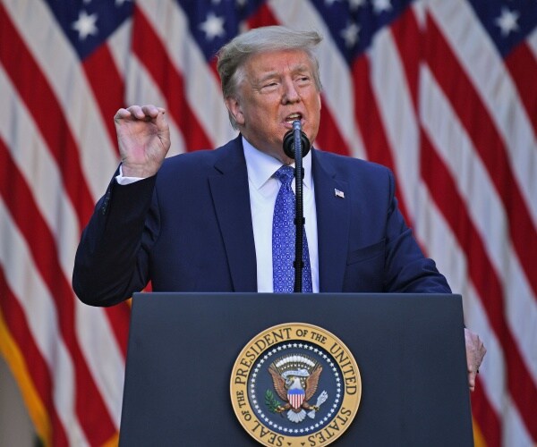 trump in a navy blue suit and lighter blue tie in front of us flags