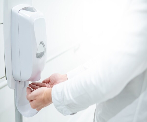 A doctor or nurse in white coat getting hand sanitizer from a dispenser in hospital hall