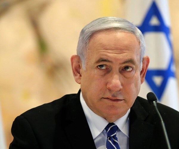 sitting in front of israeli flags, benjamin netanyahu sits in front of a mic.