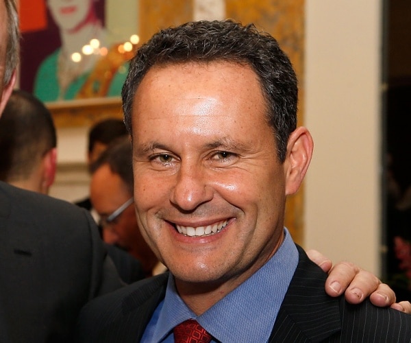 kilmeade smiling wearing a blue shirt and red tie with a black suit