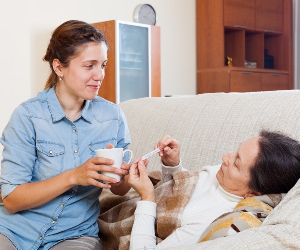 adult daughter caring for mother who is laying on couch, taking her temperature