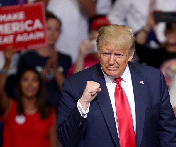 trump in a red tie and navy blue suit with supporters holding up signs behind him