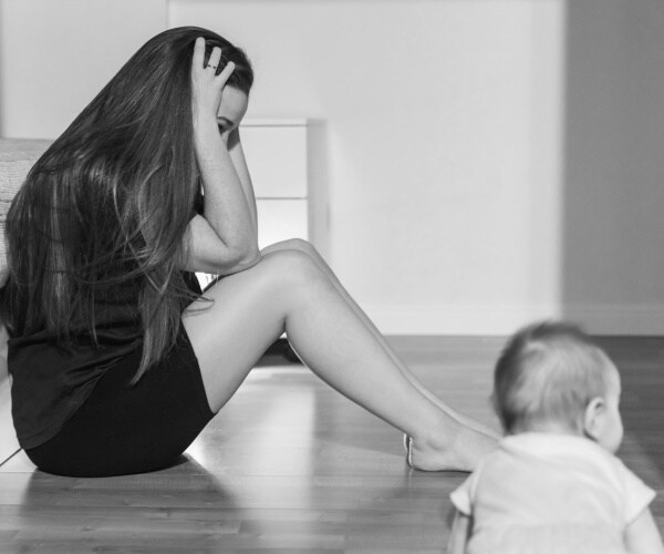 woman sitting on the floor with head in hands, baby crawling on side