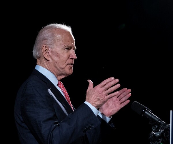 biden in a suit and red patterned tie speaking into a mic