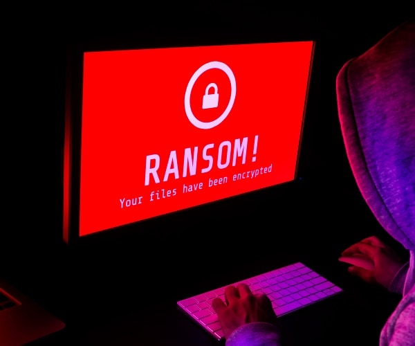 man sits in dark room at computer with red screen that says ransom