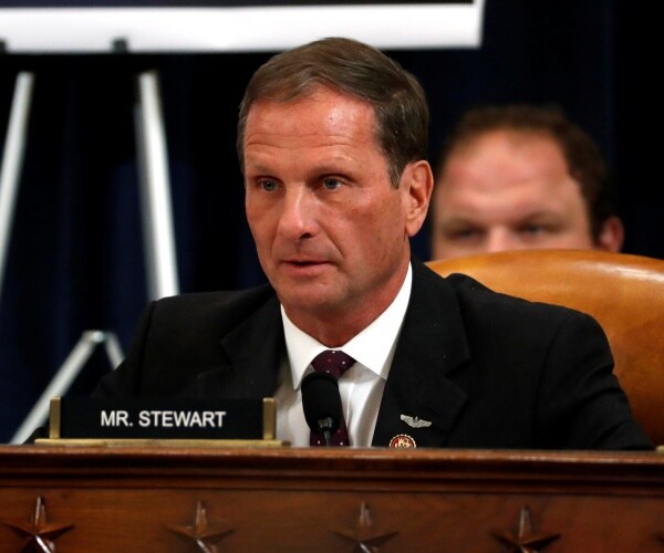 stewart in a black suit sitting at a desk with a name plate