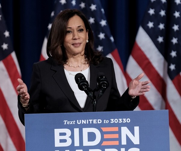 kamala harris stands at a podium with a biden-harris sign in front of flags speaking