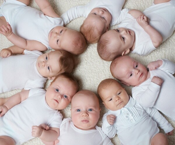 about 10 babies in a circle in white onesies, some looking up at camera