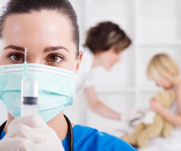 healthcare worker holds up syringe to camera, young child in background getting ready for shot