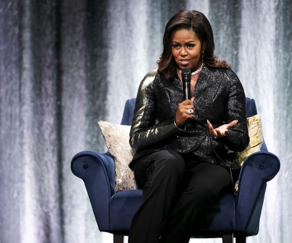 michelle obama in a shimmery black coat and black pants sitting and speaking on a stage