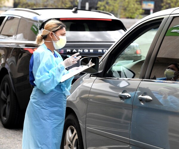 woman in blue protective suit and mask talks to a person inside a car while holding a clipboard