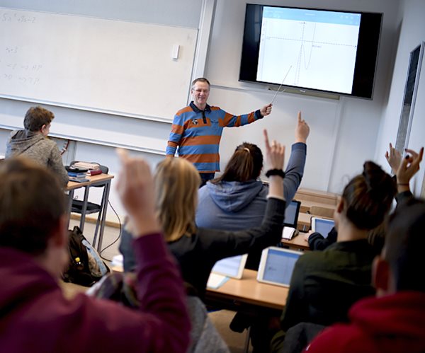 students raise their hands in a classroom while a professor points to a digital teaching tool