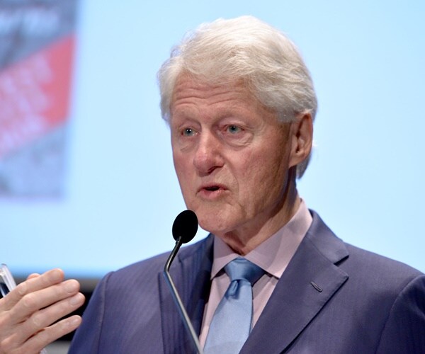 bill clinton stands in front of a mic holding glasses speaking