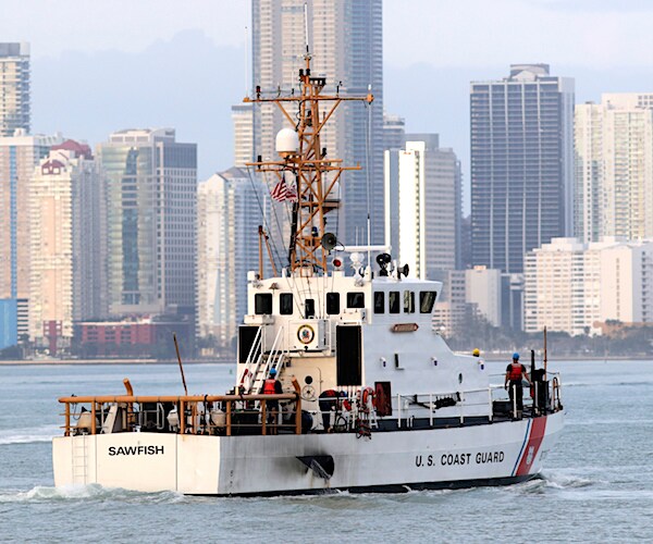 a coast guard ship floats in a bay with tall city buildings in the backdrop