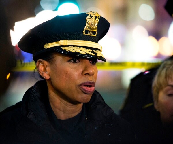 best in a police uniform and hat speaking in front of yellow caution tape and bright lights