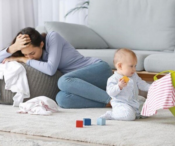 mom sitting on floor looking depressed, baby playing near her