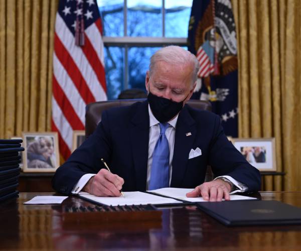 joe biden sitting at a desk signing a document with a stack of documents beside him