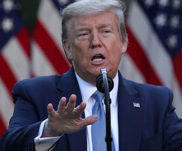 trump in a navy blue suit and light blue tie speaking in front of a us flag