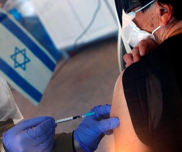 a masked woman gets inoculated by blue gloved hands with an israeli flag in the background