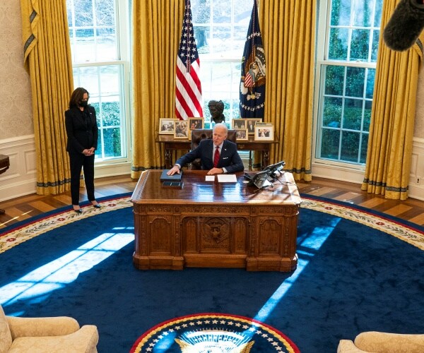 joe biden is at his desk with kamala harris nearby.