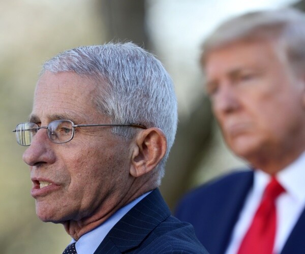 fauci in a suit and light blue shirt speaking outside with trump listening in the background