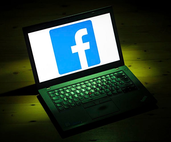 a laptop lays on a table with a large facebook logo shown on its screen in a dark room